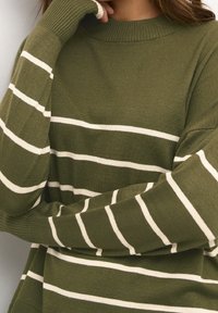Maglione lavorato a maglia verde oliva con righe orizzontali crema; presenta un colletto alto e polsini a costine. Il tessuto appare morbido e con una consistenza piacevole.