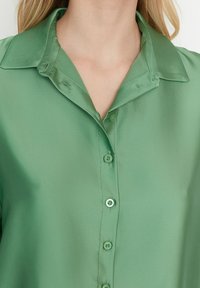 Grön satinskjorta med krage, framtida knappar och en slät yta. Har en avslappnad passform och en polerad finish.