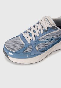 Artiškas mėlynos ir pilkos spalvų Skechers sportbačio su baltais raiščiais, tinklinėmis detalėmis ir paminkštintu padu vaizdas ant balto fono.