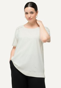 Top blanco de manga corta con escote redondo, que presenta detalles de tachuelas doradas en los hombros y un corte relajado, combinado con pantalones oscuros.