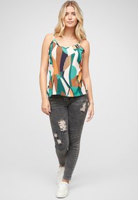 Top sin mangas multicolor con patrones abstractos en verde, naranja y crema; combinado con jeans ajustados negros desgastados y zapatillas blancas.