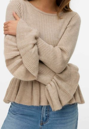 Femme portant un pull beige côtelé avec des manches évasées et un bas péplum, associé à un jean en denim bleu, posant les bras croisés.