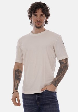 Uomo con capelli ricci che indossa una maglietta beige semplice e jeans, mostra tatuaggi dettagliati su entrambi gli avambracci, in piedi con la mano in tasca.