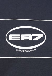 Melns tekstils ar izteiksmīgu baltu ovālu logotipu, kurā redzams "EA7" un "EMPORIO ARMANI" treknrakstā. Plānas baltas svītras izceļ dizainu.
