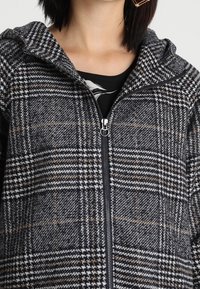 Mujer con una chaqueta de capucha de cuadros en blanco y negro, con cremallera, sobre una blusa negra, mostrando el cuello y la parte inferior de la cara.