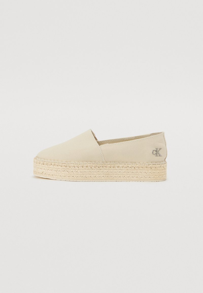 Espadrilhas em lona bege com sola de juta trançada, apresentando um bico arredondado e um detalhe de logótipo na lateral. Design clássico de colocar.