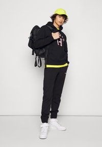 Felpa nera con logo rosso, maglietta gialla, pantaloni della tuta neri, sneaker alte bianche e cappellino giallo neon. Zaino nero.