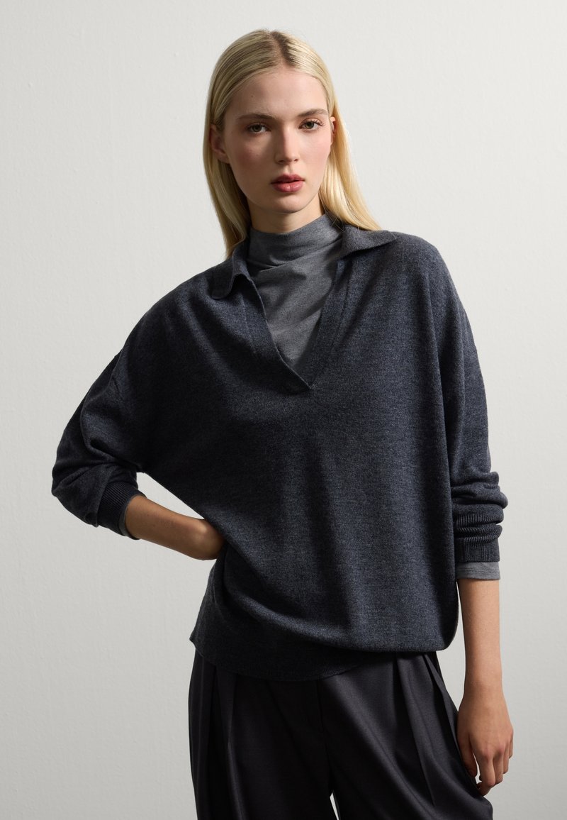 Donna bionda che indossa un maglione grigio scuro oversize sopra un dolcevita grigio e pantaloni neri larghi, in posa con una mano sul fianco contro un muro neutro.