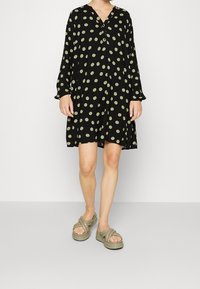 Musta mekko vihreillä pilkuilla, pitkät hihat ja napitettu V-kaula-aukko. Yhdistetty oliivinvihreisiin, paksupohjaisiin sandaaleihin.