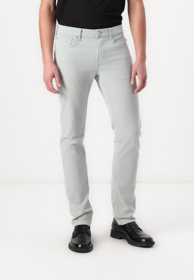 Jeans slim-fit de culoare gri deschis, cu buzunare frontale, închidere cu nasture și o textură netedă. Purtați cu pantofi de birou negri și o cămașă neagră.