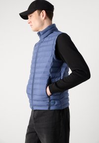 Chaleco acolchado en azul claro, con un alto cuello, cierre de cremallera y bolsillos laterales. Confeccionado en una tela suave y ligera con un ligero brillo.