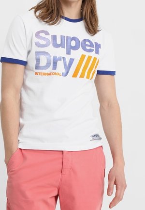Osoba ubrana w białą koszulkę z niebiesko-żółtym napisem „Super Dry International” oraz różowe szorty, ręce w kieszeniach, stojąca na białym tle.