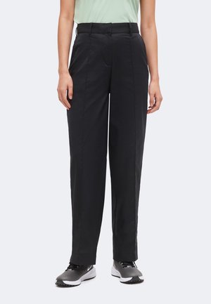 Personne portant un pantalon noir sur mesure avec des plis et des chaussures de sport noir et blanc, debout devant un fond uni.