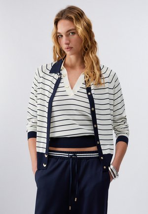 Donna che indossa un cardigan a righe bianche e blu marino sopra una maglia a V coordinata a righe e pantaloni con coulisse blu marino, in piedi con le mani nelle tasche.