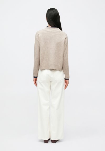 Γυναίκα με μακριά σκούρα μαλλιά, φορώντας μπεζ πουλόβερ, λευκό παντελόνι φαρδύ στα πόδια και καφέ παπούτσια, στέκεται αντικρίζοντας ένα απλό φωτεινό φόντο.