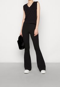 Vrouw met zwarte, mouwloze, gebreide sweater-vest, zwarte wijde broek, witte sneakers, en een zwarte bonthandtas aan de zijkant.