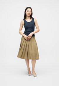 Femme portant un haut noir sans manches avec le logo "Guess", une jupe plissée beige jusqu'aux genoux, et des escarpins gris clair à bout pointu, debout les mains jointes.