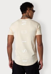 Beige Kurzarm-T-Shirt mit vertikalem weißen Text auf der Rückseite. Weiches Baumwollmaterial, abgerundeter Saum und Rundhalsausschnitt.