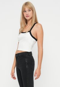 Top corto blanco con tirantes y ribetes negros; textura acanalada. Llevado con jeans negros que tienen una línea vertical de embellishments en el lateral.