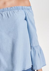 Lyseblå off-the-shoulder top lavet af glat stof, med en indsamlet elastisk halsudskæring og klokkeformede ærmer med flæsekant.