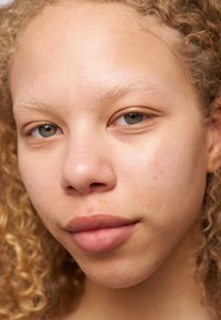 Nærbillede af et ansigt med lys hud, krøllet blond hår, gråbrune øjne, glat tekstur og naturlige læber uden makeup.