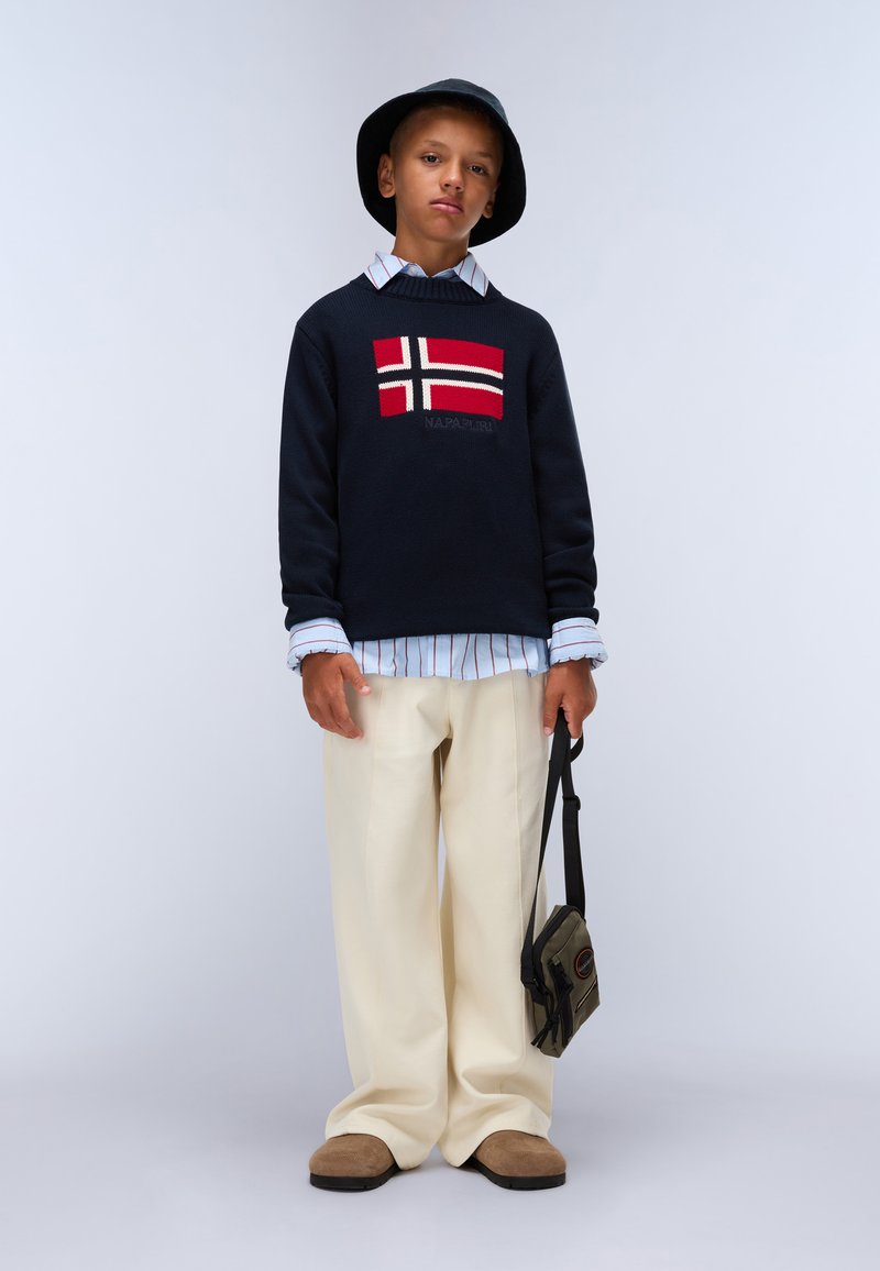 Pull marin avec drapeau norvégien tricoté, chemise à col rayée blanche, pantalon large crème, chaussures à enfiler marron et un petit sac à bandoulière gris.