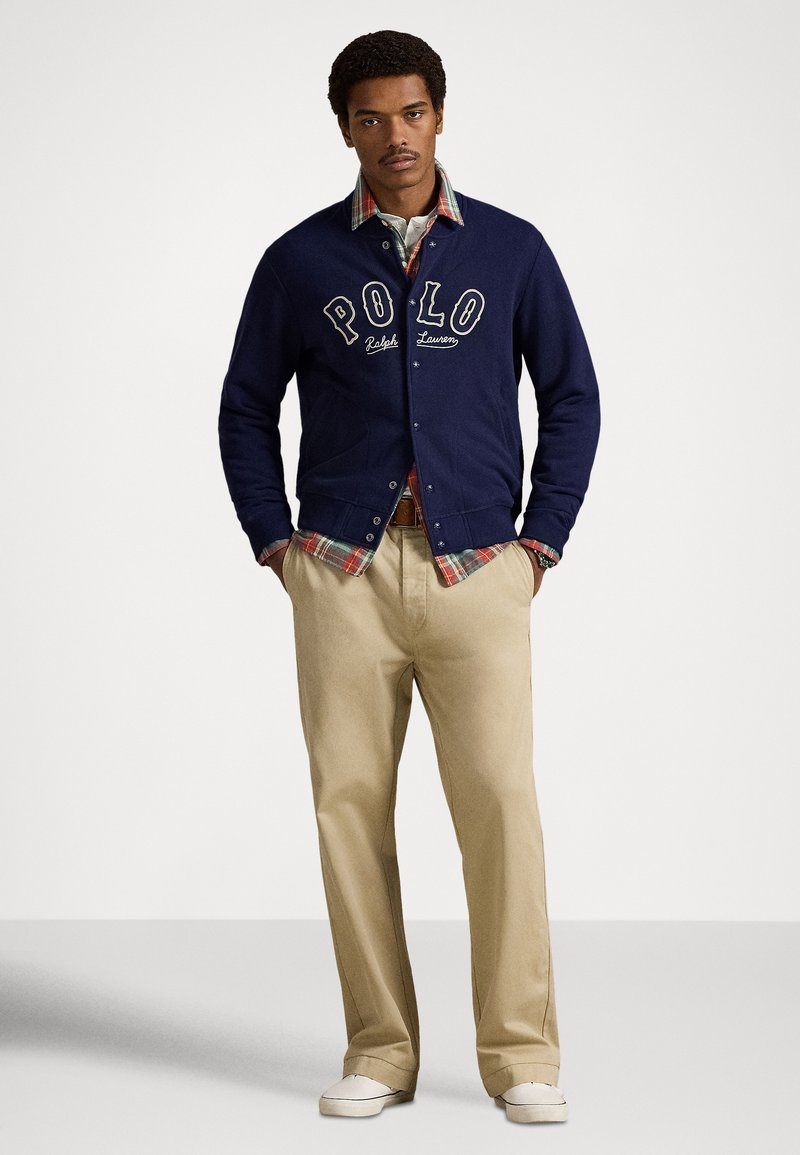 Navy cardigan with "POLO" logo, over plaid shirt, paired with tan trousers and white shoes. Casual, layered look with distinctive textures.