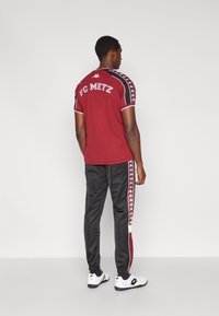 Bordowa koszulka FC Metz z czarnymi akcentami, krótkimi rękawami i białym wykończeniem; w połączeniu z czarnymi spodniami z czerwono-białymi paskami po bokach.