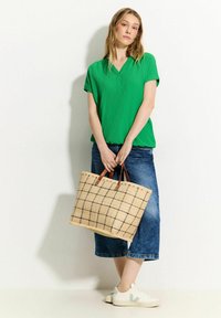 Top verde ricamato con scollo a V, abbinato a una gonna di jeans. Tiene in mano una borsa a tracolla intrecciata con manici marroni e un motivo a griglia. Indossa sneakers bianche.