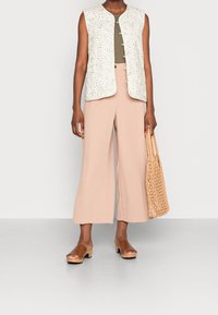 Chaleco blanco sin mangas con un patrón floral, top verde oliva, pantalones de pierna ancha rosa pálido, zuecos marrones y bolso de paja trenzada en la mano.