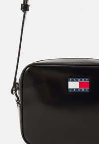 Bolso bandolera de charol negro con forma rectangular, que presenta un logo de Tommy Jeans en rojo, blanco y azul en la parte frontal.