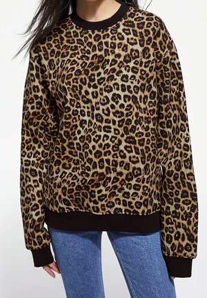 Mujer con un jersey de manga larga con estampado de leopardo, puños y bajo negros, combinado con vaqueros azules, sobre un fondo liso.