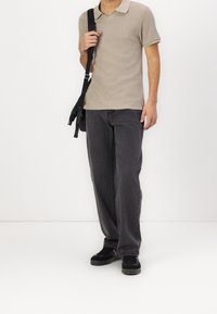 Polo beige à manches courtes avec un motif texturé, associé à un jean gris foncé ample et des chaussures noires. Modèle tenant un sac noir.