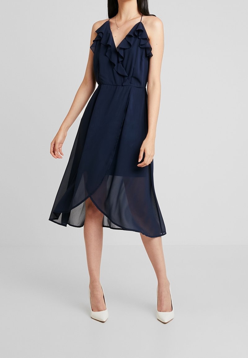 Robe midi bleu marine avec encolure à volants et fines bretelles, présentant un ourlet diagonal et une superposition transparente. Portée avec des talons blancs pointus.