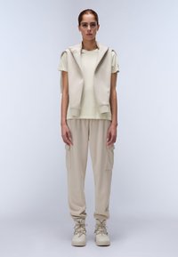 Felpa senza maniche beige sopra una maglietta verde chiaro, abbinata a pantaloni cargo beige e stivali con lacci beige. Tessuto morbido, vestibilità rilassata.