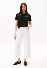 Fekete pamut póló fehér logóval, fehér bő szárú farmerrel és fekete slip-on cipővel párosítva. A ruházat minimalista dizájnt képvisel.