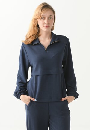 Femme aux cheveux blonds portant un haut à manches longues bleu marine avec un col demi-fermeture éclair et un pantalon assorti, mains dans les poches, expression neutre.