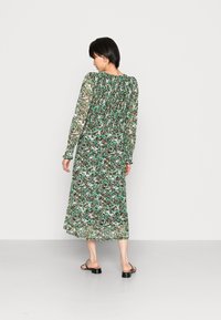 Vestido floral de manga longa em tecido verde translúcido, com costas franzidas, apresentando uma silhueta fluida e sandálias pretas discretas.