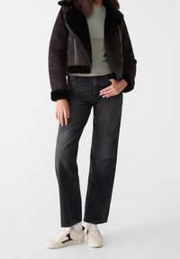 Svart shearlingjacka med bred krage, grön åtsittande skjorta under, högmidjade svarta jeans och beige sneakers med svarta accenter.