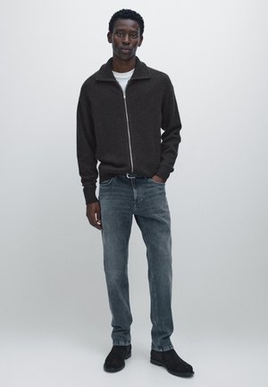 Maglione marrone con zip e colletto alto, abbinato a jeans grigi e scarpe nere. Il maglione presenta un tessuto strutturato e una vestibilità comoda.