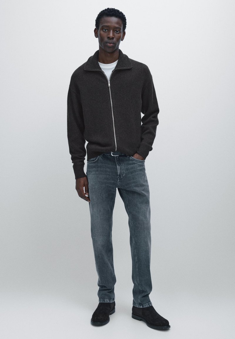 Brown zip-up sweater with a high collar, paired with grey jeans and black shoes. The sweater has a textured fabric and relaxed fit.