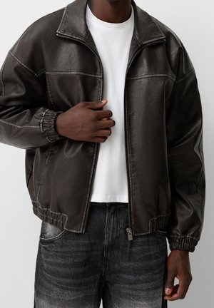 Person wearing an open dark brown leather jacket over a white shirt and faded black jeans, with one hand holding the jacket lapel.