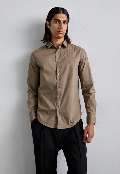 Chemise marron à manches longues avec un col pointu, fermeture à boutons, et un petit logo sur la poitrine, associée à un pantalon noir ample.