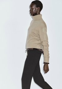 Maglione beige a collo alto con polsini a costine, abbinato a pantaloni neri. Il modello è in piedi di lato su uno sfondo neutro, mostrando l'outfit.