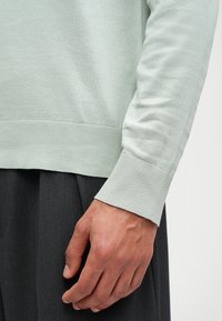 Světle zelený svetr z měkkého, hladkého materiálu s žebrovanými manžetami a lemem. Ruka, která je viditelná, jemně spočívá na tmavě šedých kalhotách.