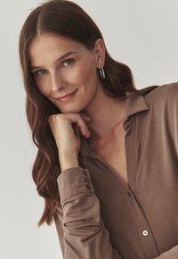 Camicia marrone a maniche lunghe con chiusura a bottoni e texture morbida. Il modello poggia la mano sotto il mento, mostrando orecchini a cerchio in argento.