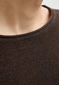 Nærbillede af en persons nakke og øvre bryst iført en mørkebrun tekstureret striktrøje med en rund halsudskæring.