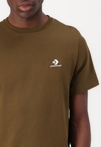 Camiseta de algodón verde oliva con cuello redondo, mangas cortas y un logo blanco de Converse en la parte izquierda del pecho. Textura suave.