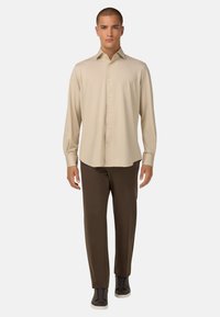 Camicia beige a maniche lunghe con colletto a bottoni, abbinata a pantaloni marroni e sneaker scuri. Vestibilità slim con texture del tessuto liscia.