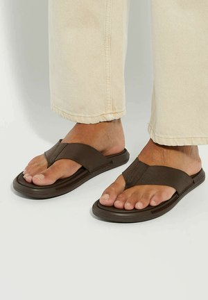 Brown sandals with a flip-flop design, featuring a wide strap made from textured material and a flat sole, worn with light-colored pants.